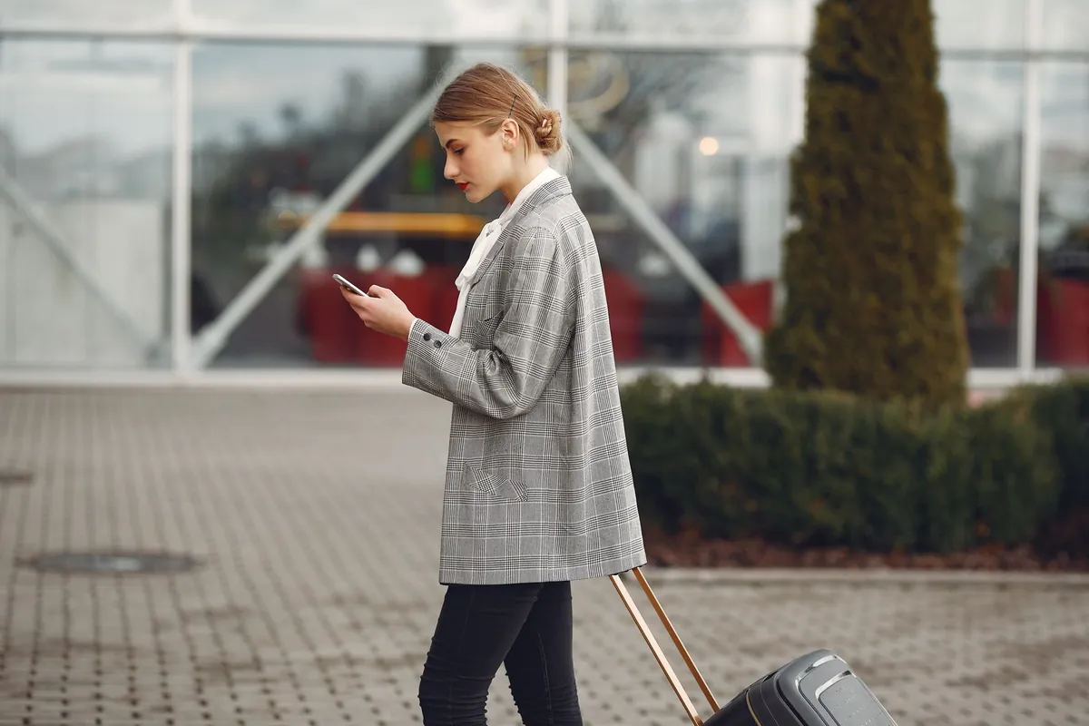 Reiziger met koffer kijkt op haar telefoon bij de ingang van een vliegveld
