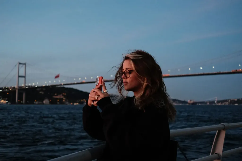Vrouw maakt met haar telefoon een foto bij de Bosporus in Istanbul, met de Bosporusbrug op de achtergrond
