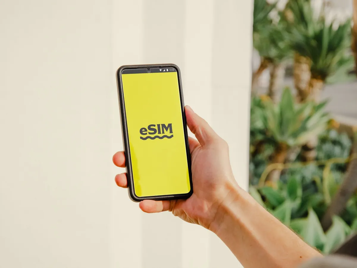 Hand houdt een smartphone vast met het woord eSIM op een geel scherm, buiten tussen planten