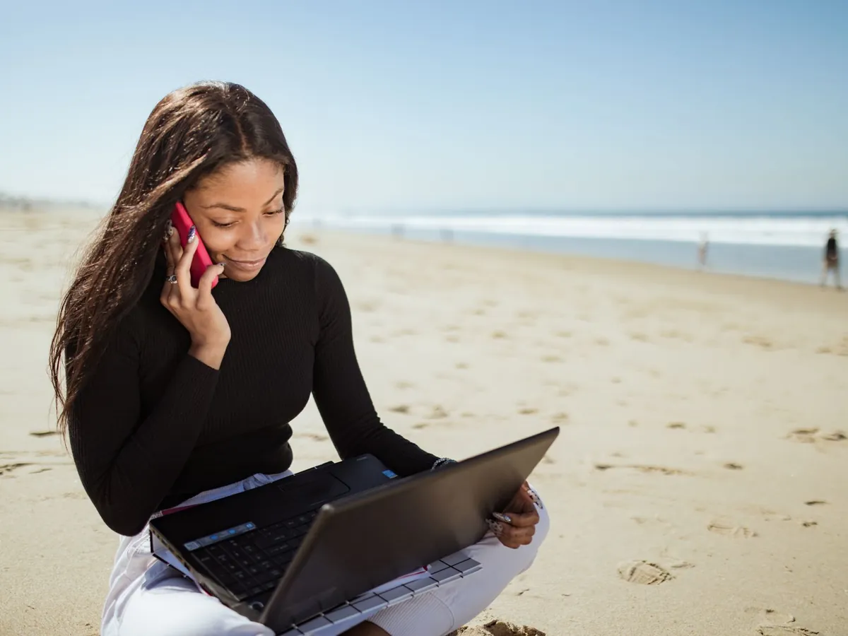 Vrouw zit op het strand met een laptop op schoot en belt met haar smartphone
