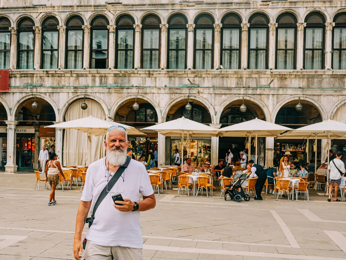 Man met schoudertas staat op een zonnig Piazza San Marco in Venetië met terrassen op de achtergrond